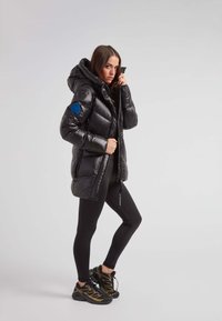 Czarna kurtka puchowa o błyszczącym wykończeniu, z kapturem i niebieską naszywką, noszona na czarne legginsy i buty sportowe.