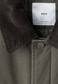 Giacca verde scuro con colletto in velluto a coste nero, bottone a pressione metallico con il testo "King Denim" e etichetta con taglia MNG all'interno del colletto.