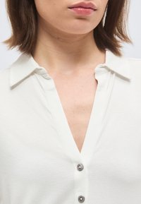 Femeie purtând o cămașă albă cu guler și două nasturi vizibili, arătând o parte din gât, bărbie și buze pe un fundal simplu.