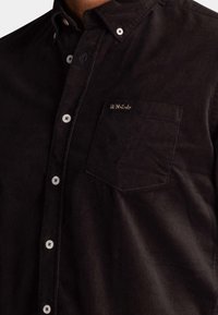 Zwarte corduroy shirt met een button-down kraag, witte knopen en een borstzak met geborduurde tekst: "*N.Z.A.*" in goud.