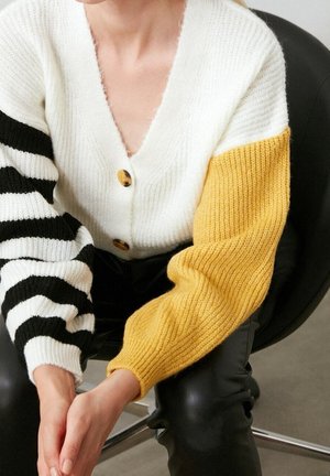 Personne portant un cardigan blanc aux manches rayées jaunes et noires, assise les mains jointes sur un pantalon en cuir noir et sur une chaise noire.