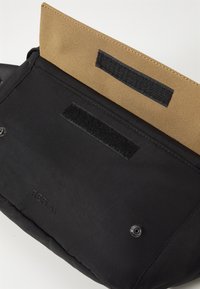 Black bag with a textured outer material, featuring a tan fabric flap secured by two black Velcro strips and a logo embossed on the front.