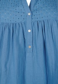 Blouse bleu clair avec un col en V, un bas plissé et des coutures décoratives sur le corsage. Elle est ornée de boutons rappelant des perles sur le devant.