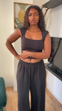 Mulher com cabelo encaracolado vestindo um top cropped preto e calças largas está dentro de casa, com a mão na cintura, com uma obra de arte de abacaxi e uma televisão ao fundo.