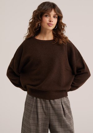 Femme aux cheveux bruns ondulés portant des boucles d'oreilles anneaux argentées, un pull ample marron foncé et un pantalon à carreaux gris taille haute, debout devant un fond uni.