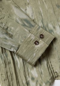 Camicia verde con motivo a fantasia e texture stropicciata, con bottoni marroni ai polsini e un design fluido.