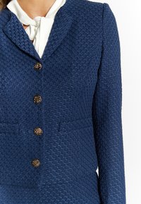 Blazer navy con texture e design aderente, dotato di colletto a scollo a V, sei chiusure decorative con bottoni dorati e due tasche frontali.