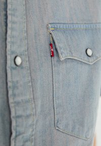 Camicia di jeans azzurro chiaro con taschino frontale. Presenta cuciture gialle in contrasto e un bottone a pressione nero. Etichetta del marchio rossa visibile.
