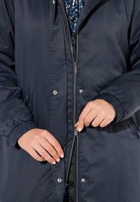 Marinblå zip-up jacka med en slät textur, elastiska ärmmuddar och tryckknappar. Har en sidoficka och kontrasterande innertyg.