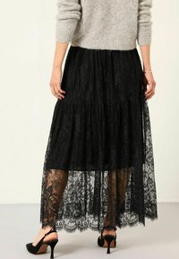 Schwarzer Spitzen-Maxi-Rock mit gestuftem Design, transparenter Textur, gewelltem Saum, kombiniert mit schwarzen High-Heels. Oben ein Wollpullover.