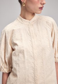 Helles beiges Blusenshirt mit floralem Stickerei, hohem Kragen und gepufften Ärmeln mit elastischen Bündchen. Knopfverschluss vorne.
