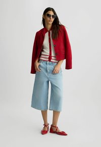 Jachetă roșie din suede, bluză albă din tricot cu accente roșii, blugi evazați de culoare albastru deschis și pantofi plan roșii. Modelul poartă ochelari de soare negri.