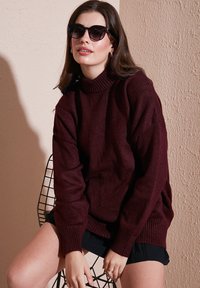 Burgundi kötött pulóver magas nyakkal és bordázott mandzsettával, sötét napszemüveggel párosítva. A modell egy fekete drótszéken ül egy texturált fal előtt.