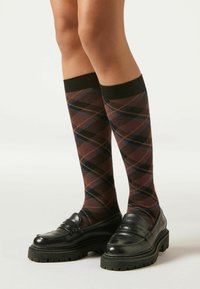 Knielange sokken in bruin met een ruitpatroon in blauw en zwart, gedragen met zwarte loafers met een dikke, gestructureerde zool.