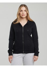 Tânără cu păr blond lung, purtând un hanorac negru cu fermoar și pantaloni de trening gri deschis, stând pe un fundal simplu.