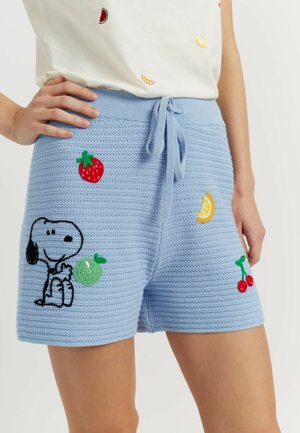 Shorts en maille bleu clair avec cordon, présentant un Snoopy brodé et divers patchs de fruits, portés par une personne la main sur la hanche.