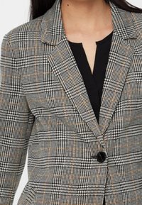 Šedý kostkovaný blazer s černými a oranžovými detaily, strukturovaný design s vykrojeným límcem, zapínání na jeden knoflík a hladká textura.