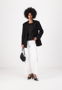 Blazer nero con rever, indossato sopra una maglia scura. Jeans bianchi a gamba larga, scarpe nere con tacco e una piccola borsa nera.