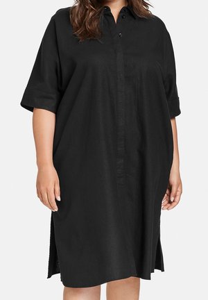 Woman wearing a loose-fitting black knee-length shirt dress with short sleeves and a buttoned collar.