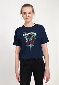 Mladá žena s drdolem, oblečená v tmavě modrém tričku, na kterém jsou barevná sluchátka a nápis "STRAIGHTFIRE Tropical Twist."