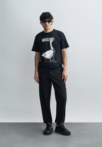 Čierne tričko s bielou grafikou husi, doplnené o čierne nohavice a čierne tenisky. Na tričku je nápis "MOSCHINO".
