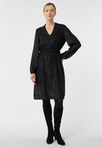 Czarna sukienka z długimi, przezroczystymi, paskowanymi rękawami, dekoltem w kształcie litery V i talią podkreśloną paskiem. Noszona z czarnymi botkami za kolano.