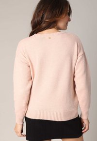 Pull gris rose surdimensionné en tissu doux, doté d'un ourlet et de poignets côtelés, avec un petit accent en forme de cœur doré au niveau du col.