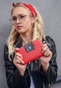 Red wallet made of textured material, with a black polka-dotted flap and a metal snap button. Features a silver logo on the front.