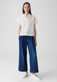 Blusa bianca in pizzo con maniche corte, apertura con bottoni; abbinata a pantaloni in denim scuro a gamba larga e sneakers bianche, che mostrano una vestibilità rilassata.