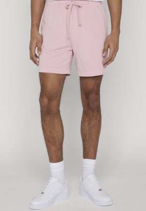 Mężczyzna w jasnoróżowych szortach ze sznurkiem, białych skarpetach sięgających do kostek i białych trampkach, stojący na tle jednolitego jasnego tła.