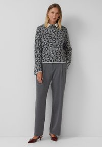 Vrouw in een grijze sweater met luipaardprint, grijze streepjesbroek, wit coll shirt en donkerrode puntige hakken, staande tegen een grijze muur.