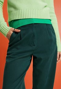 Zelený žebrovaný svetr přes zelenou košili, doplněný tmavě tyrkysovými kalhotami s předními sklady a bočními kapsami, umístěný na oranžovém pozadí.