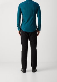 Turquoise overhemd met knopen, lange mouwen, nauwsluitend ontwerp en een afgeronde zoom, gecombineerd met zwarte jeans met rechte pijpen en zwarte schoenen.