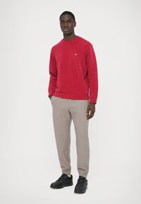 Röd quiltad sweatshirt med rund hals och liten logga på bröstet, kombinerad med ljusgrå texturerade byxor och svarta sneakers.