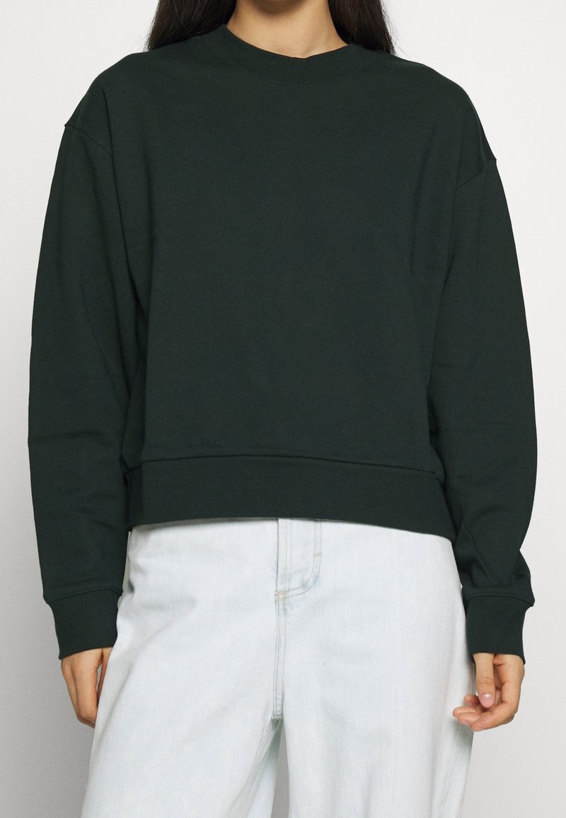 Personne portant un sweat-shirt à manches longues vert foncé et un pantalon blanc taille haute, debout devant un fond uni.