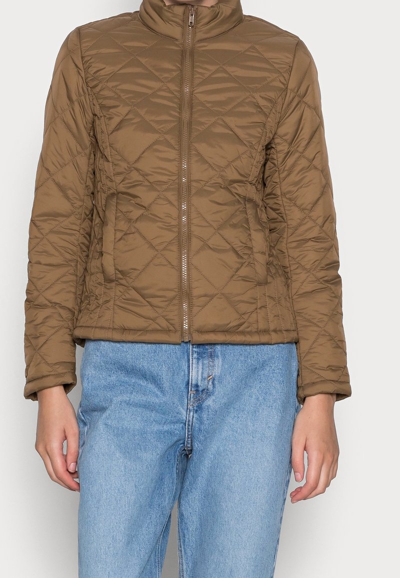 Personne portant une veste matelassée marron avec une fermeture éclair à l'avant et un jean en denim bleu, debout devant un fond uni.