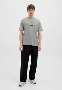 Grey cotton t-shirt with a round neckline and short sleeves, featuring a black logo print on the chest. Paired with loose black pants and sneakers.