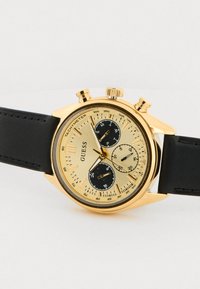 Zlaté hodinky s textúrovaným ciferníkom, čiernymi subciferníkmi a čiernym koženým remienkom. Funkcie zahŕňajú tachymeter a chronograf.