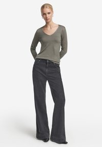 Olivgrön långärmad topp med v-ringning, som är ihopparad med högmidjade svarta utsvängda jeans. Tyget verkar mjukt med en slät yta.