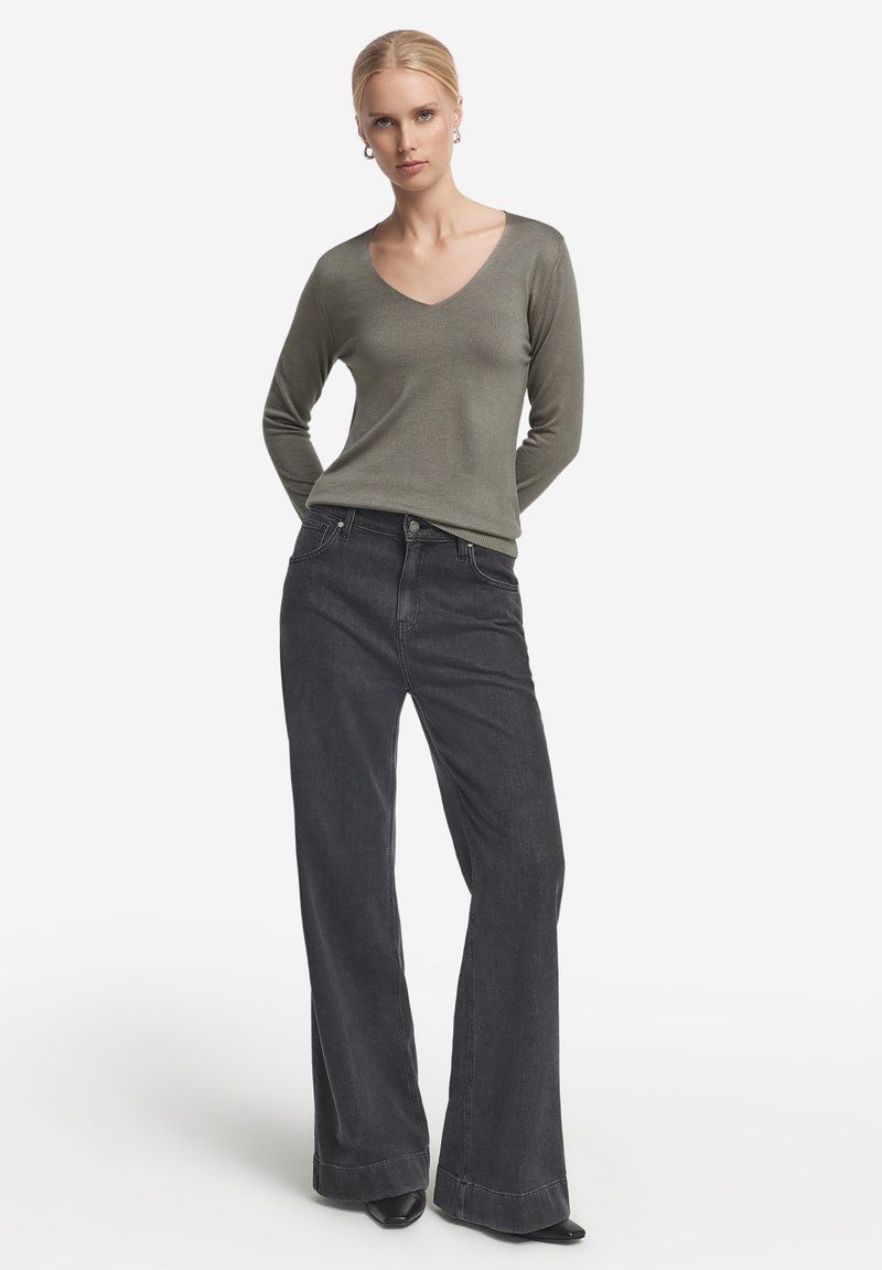 Olivgrön långärmad topp med v-ringning, som är ihopparad med högmidjade svarta utsvängda jeans. Tyget verkar mjukt med en slät yta.