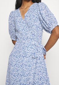 Blaues Blumenkleid mit Puffärmeln, V-Ausschnitt, geraffter Taille und Bindedetail. Mit einem dichten Muster aus weißen und dunkelblauen Blumen.