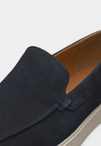 Chaussure à enfiler en daim bleu marine avec doublure intérieure beige et semelle en caoutchouc blanche, présentant des coutures détaillées le long des bords supérieurs et latéraux.