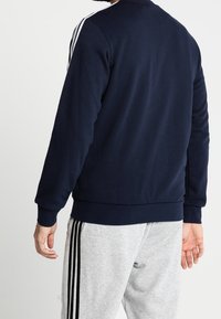 Sudadera azul marino de manga larga, con puños y dobladillo acanalados. Presenta acentos de tres franjas blancas en los hombros. Textura suave.