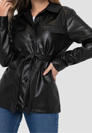 Femme portant une veste ceinturée en fausse cuir noire avec des poches à boutons et un jean bleu sur un fond uni.