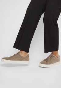 Világos bézs velúr sneaker, fehér gumi talppal, kerek orral, fűzős dizájnnal és minimális varrási részletekkel. Fekete nadrággal párosítva.