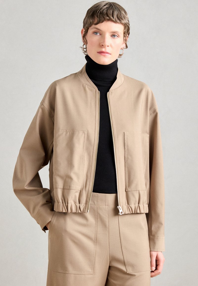 Beige kort jacka med dragkedja fram, två framfickor och elastisk midja; bärs över en svart ribbad turtleneck. Slät textur.