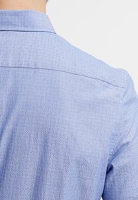 Vaaleansininen paita, jossa on teksturoitu pilkkukuvio, klassinen kaulus ja sileä kangas. Näkyvät saumat ja siisti takaosa.