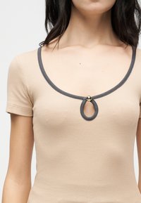 Γυναίκα που φοράει μπεζ ριγέ τοπ με κοντά μανίκια και στρογγυλή λαιμόκοψη με σκούρες γκρι λεπτομέρειες, στολισμένο με άνοιγμα σε σχήμα σταγόνας και χρυσό κουμπί.