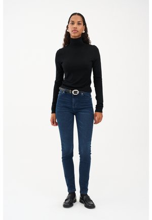 Ženska z rdečim puličastim puloverjem, temno modrimi kavbojkami z pasom, črnimi loaferji, stoji pred enobarvnim belim ozadjem.