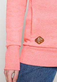 Korallfärgad sweatshirt med ribbade ärmslut och nederkant, tillverkad av mjukt material. Har en brun logopatch på den nedre sidan.